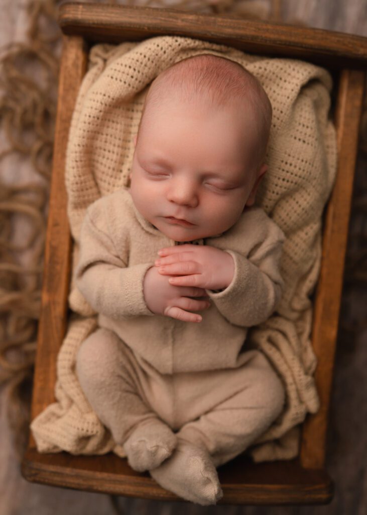 Sleeping newborn baby boy in a tan outfit resting on textured blankets inside a wooden bed prop.