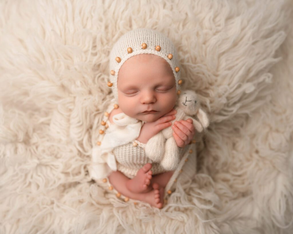 Newborn baby boy wrapped in cream knit with a beaded bonnet while holding a soft cream teddy bear.