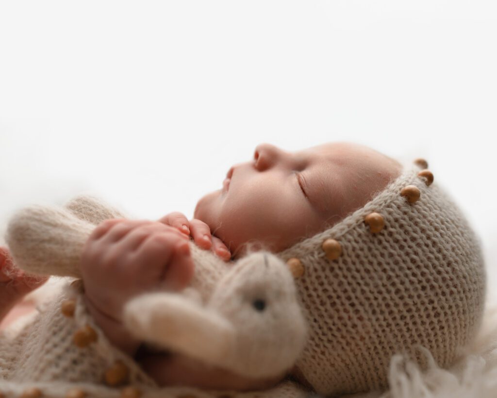 Sleeping baby boy holding a white bunny while wearing a cream knit bonnet with wooden bead accents.
