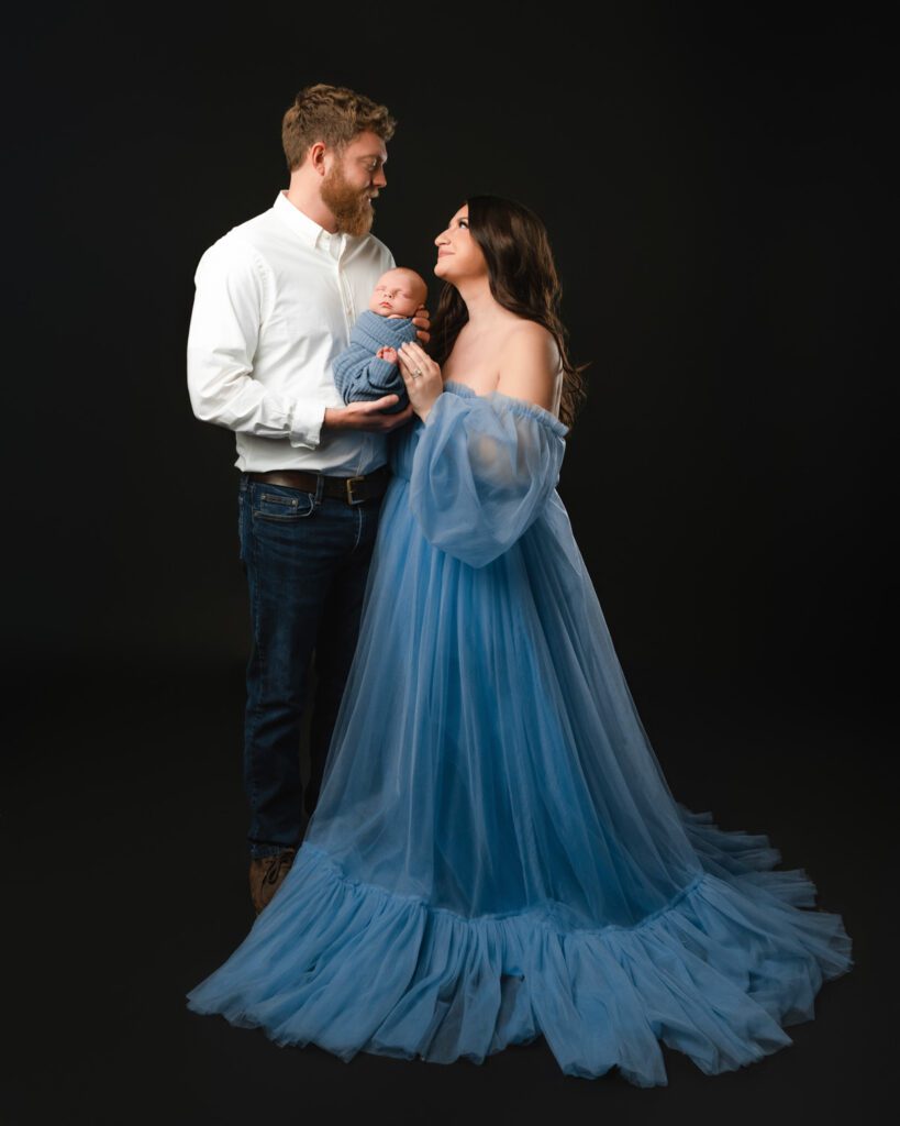 Parents holding their newborn baby boy against a black backdrop, with mom wearing a flowing blue tulle dress.