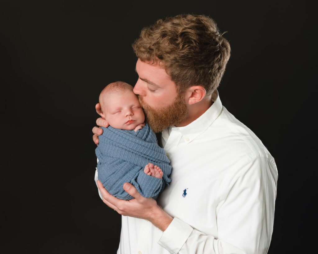 Dad in a white shirt holding his swaddled newborn son close while kissing his cheek.