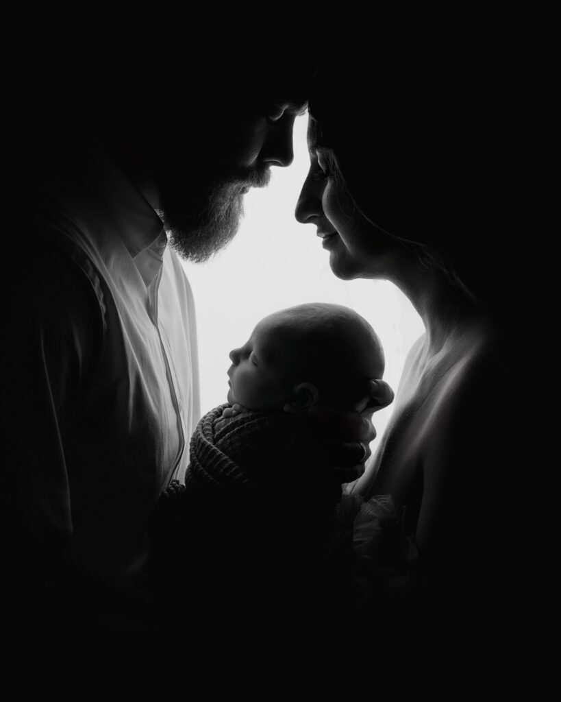 Black and white silhouette of parents facing each other while holding their newborn baby between them.