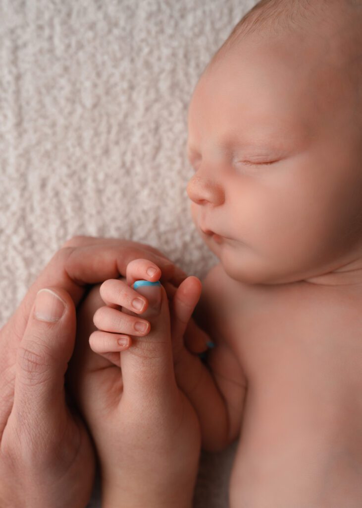 Parents gently holding their newborn baby boy’s hand as he sleeps on a soft cream blanket.