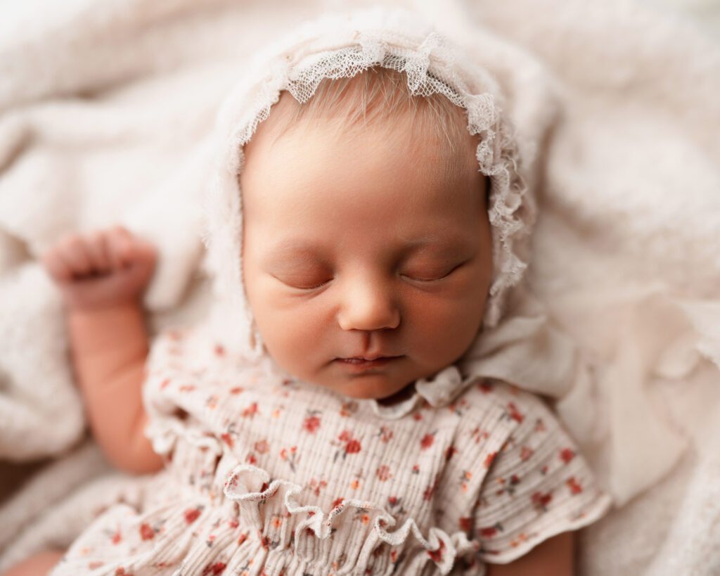 Close-up of a sleeping newborn baby girl in a light pink bonnet and floral outfit on a soft cream backdrop.