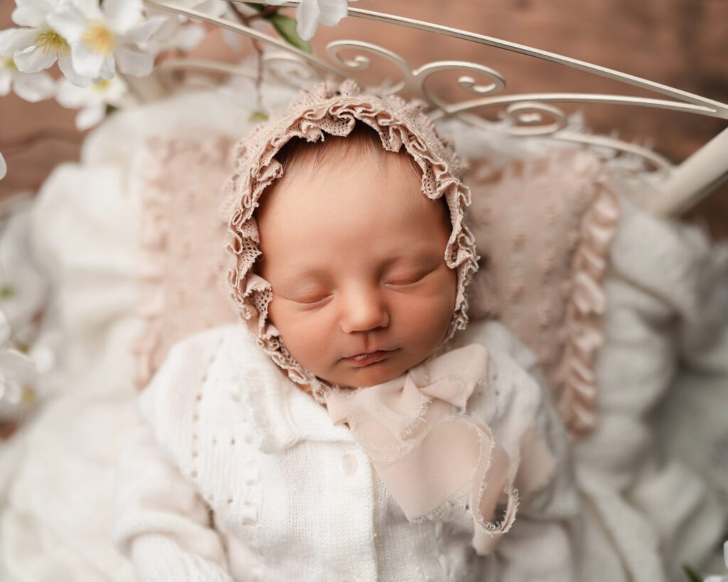 Sleeping newborn baby girl in a pink ruffled bonnet and white knit sweater resting on textured cream blankets.