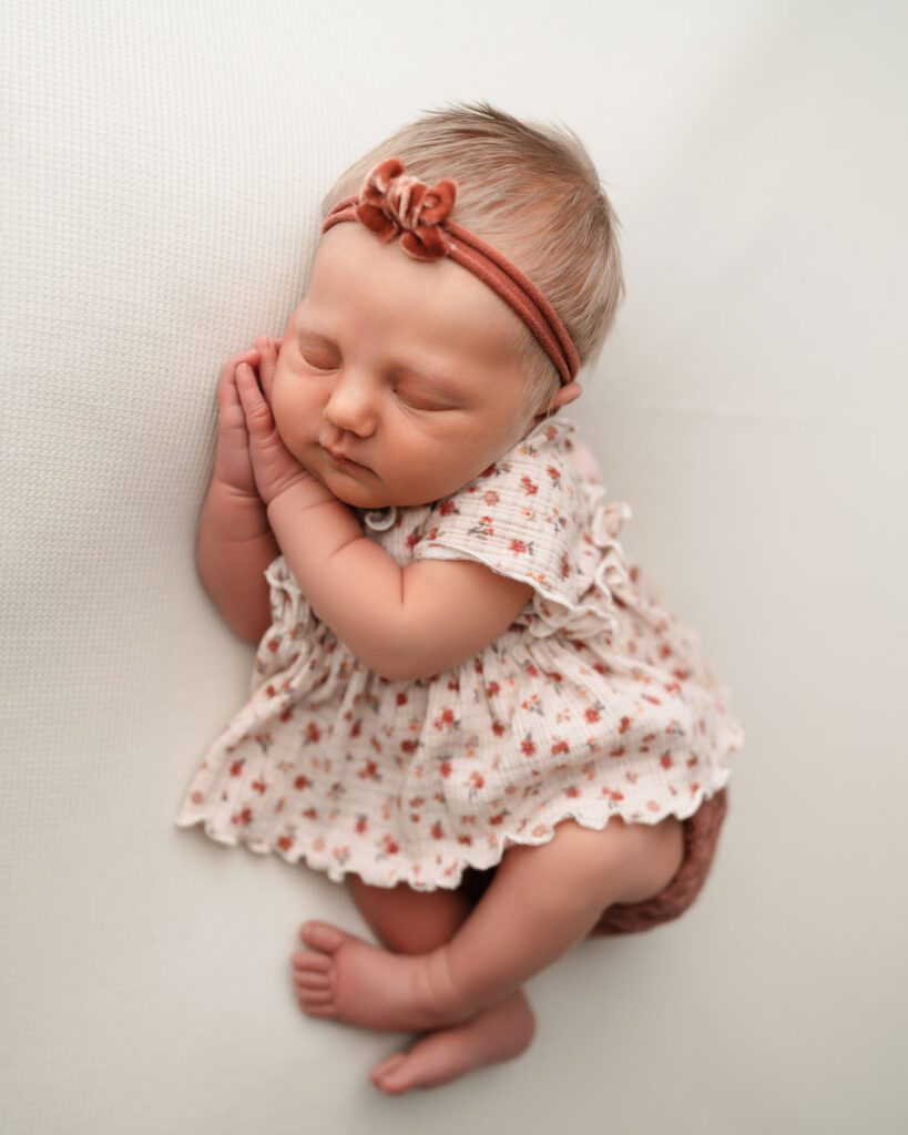 newborn baby girl in vintage floral outfits and bow headband laying on her side with hands under her cheek.