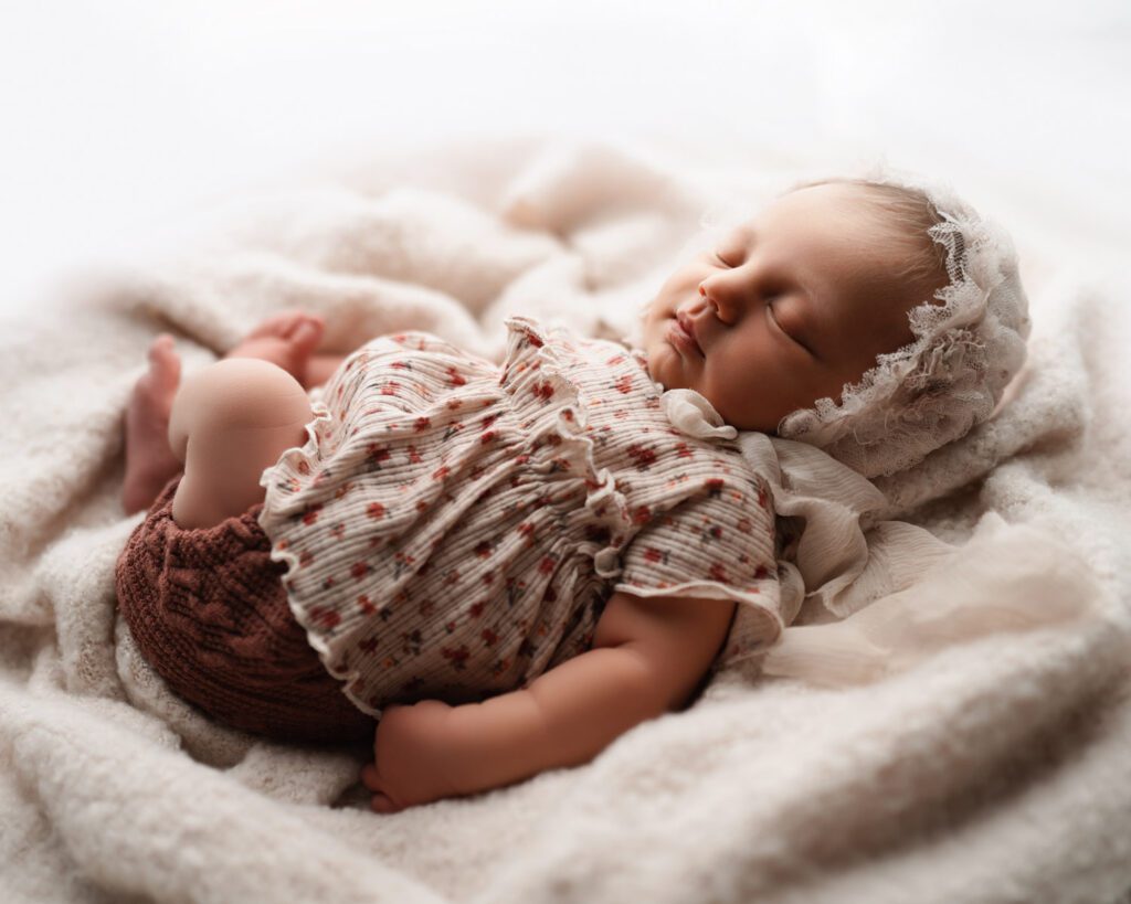 Newborn baby girl sleeping on a plush cream blanket with soft window light across her face.