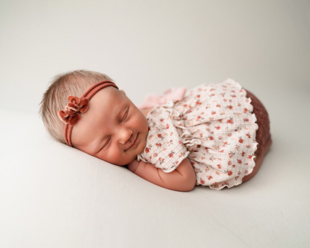 Smiling newborn baby girl in a pink floral outfit and rust bow headband sleeping on a clean white backdrop.