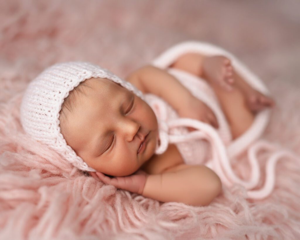 sleeping newborn girl swaddled in pink knit wrap and bonnet laying on her side on pink flokati rug