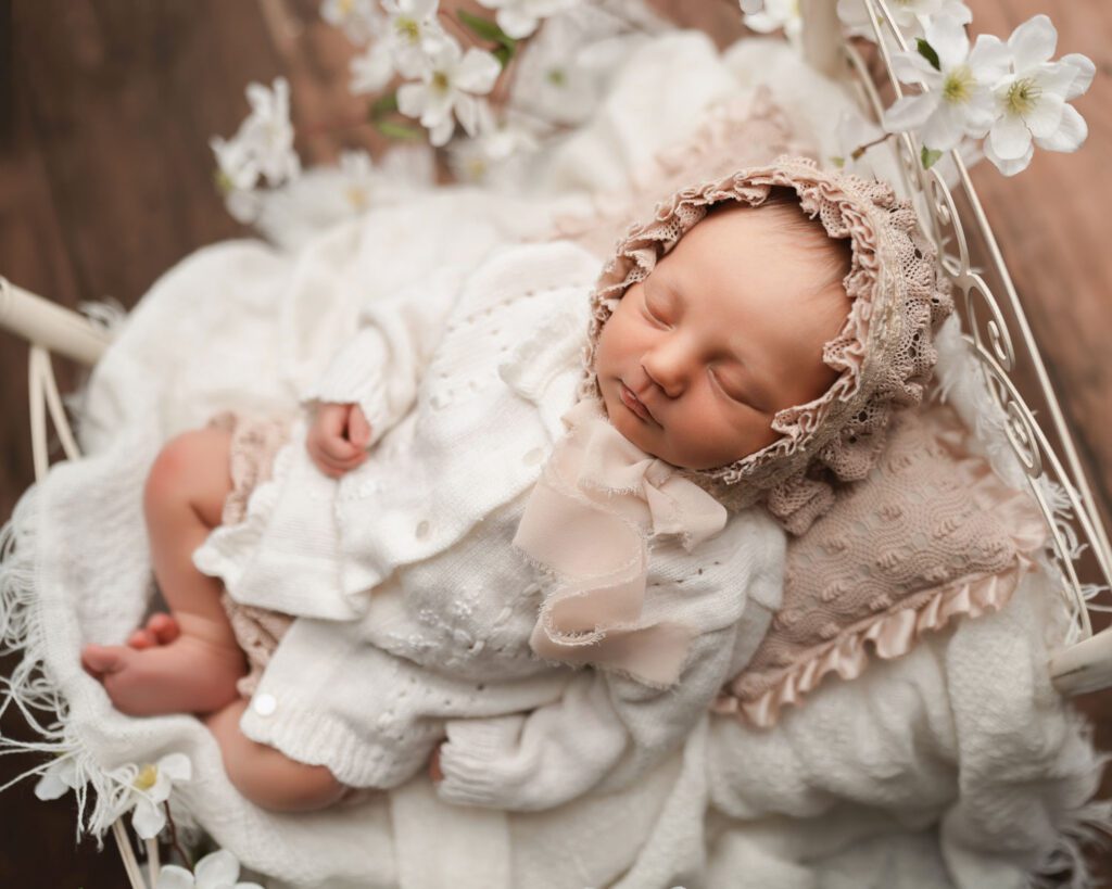 Newborn baby girl sleeping on a white iron bed with vintage blankets and white flowers surrounding bed.
