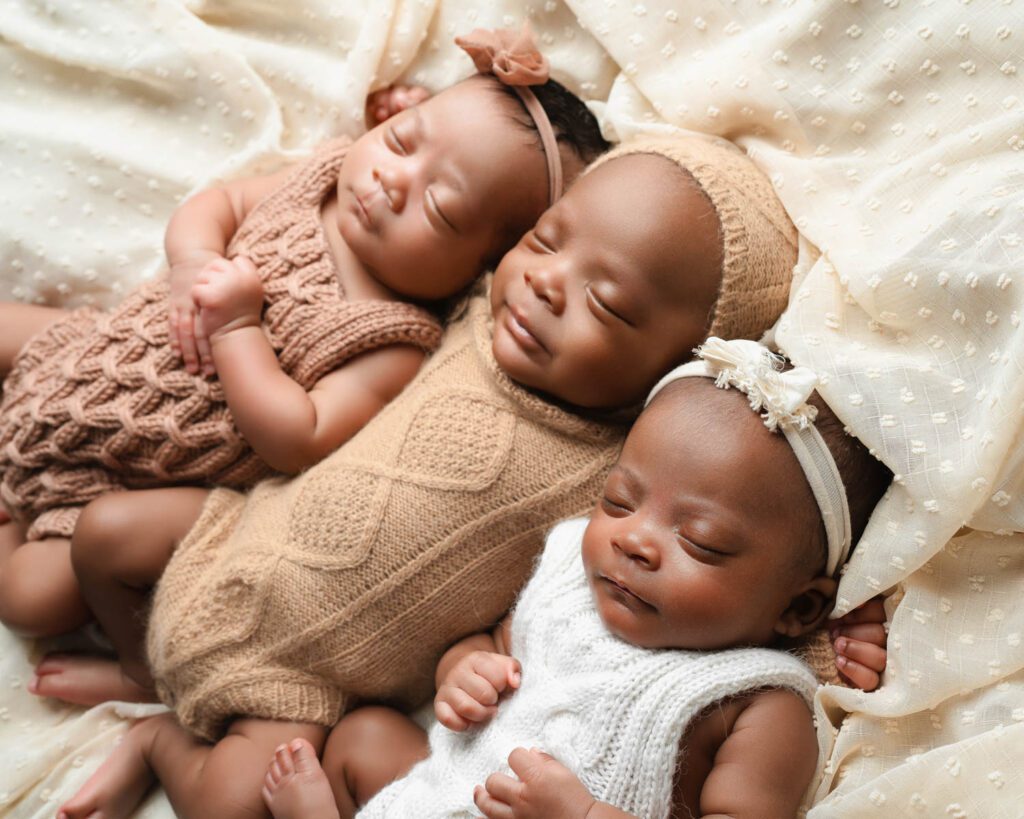 Three newborn triplets sleeping side by side in neutral knit rompers on a soft cream blanket.