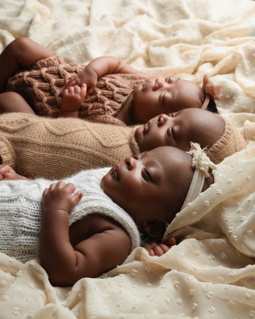 Three newborn babies resting side by side on a cream blanket during a triplets newborn photos session.