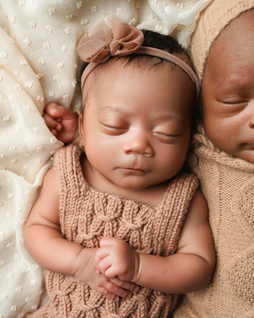 Baby girl in a brown knit romper holding her hands together while lying beside her brother.