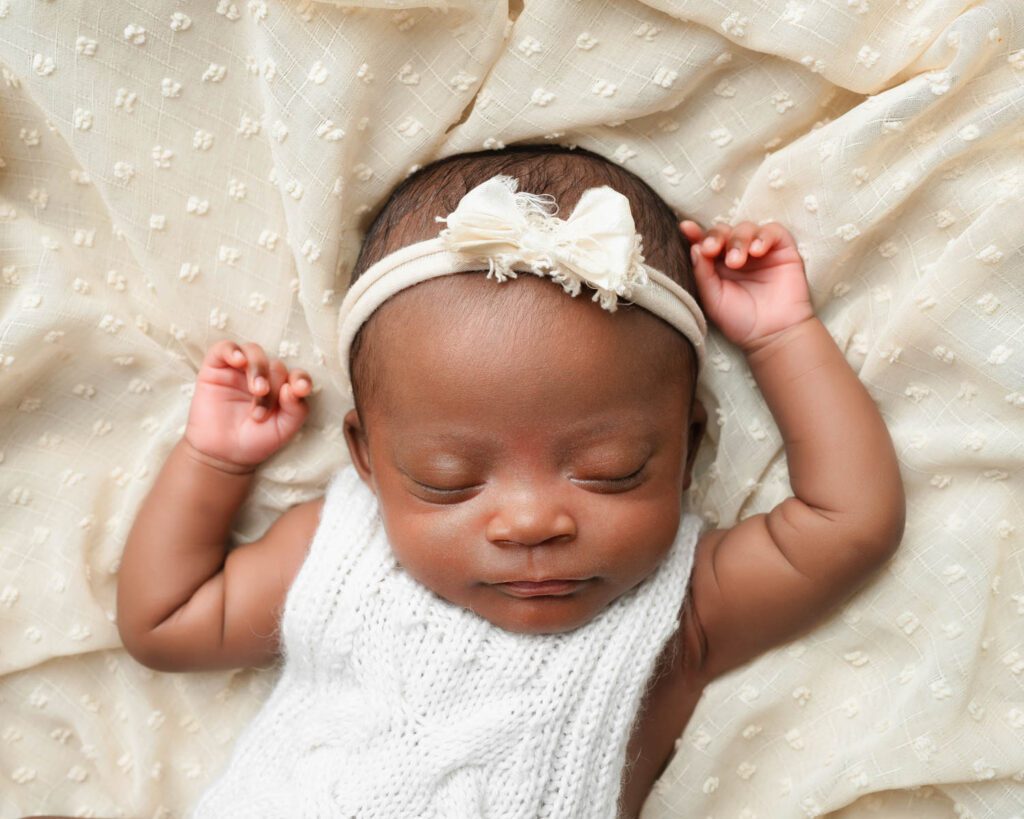 Sleeping newborn baby girl in a white knit romper and bow headband with both arms raised on a cream blanket.