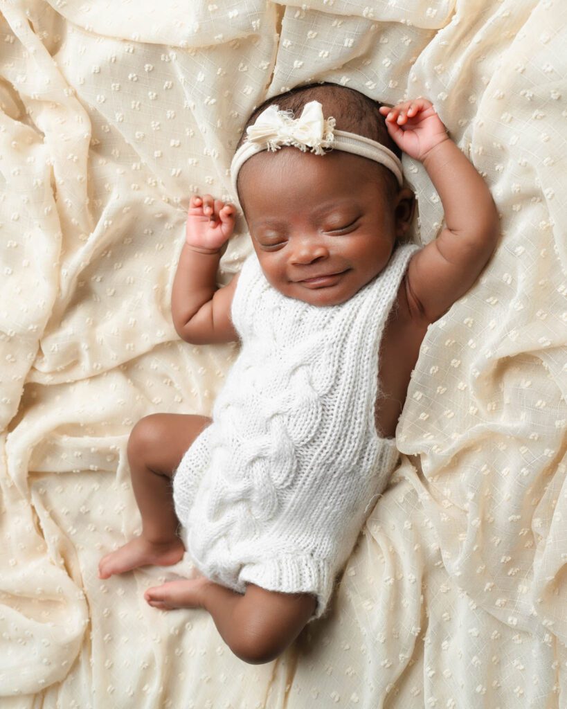 Newborn baby girl smiling in her sleep while wearing a white knit romper and floral bow headband.