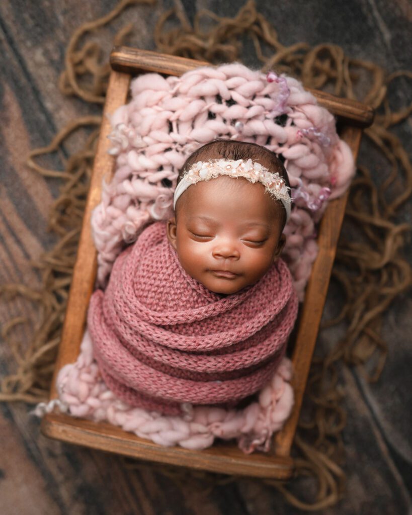 Sleeping newborn baby wrapped in dusty pink knit fabric with a matching bonnet in a wooden crate.