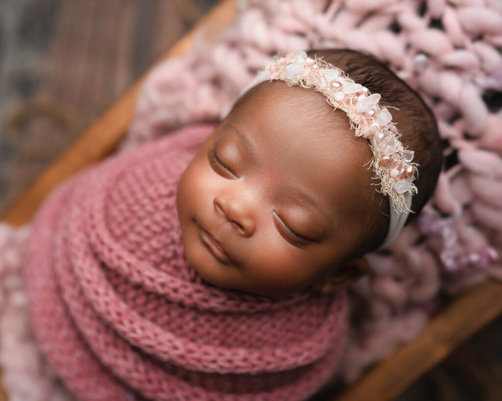 Baby girl wrapped in pink with a beaded headband while sleeping in a wooden crate.