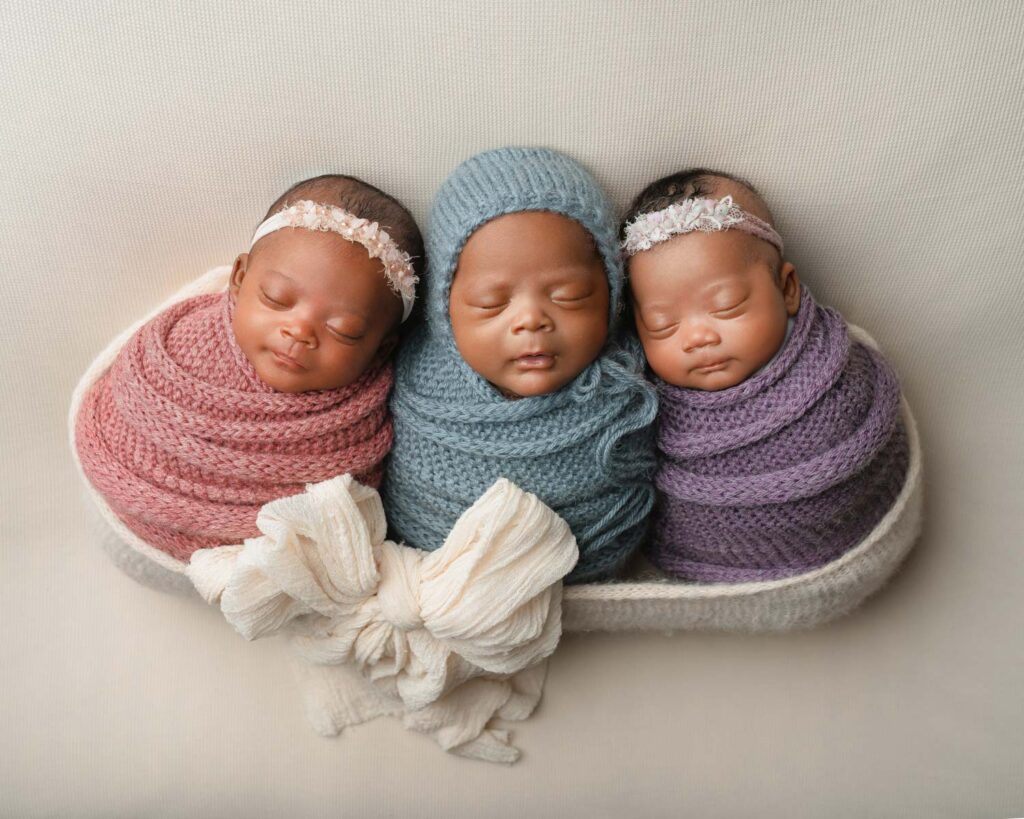 Three newborn triplets swaddled in pink, blue, and purple wraps while sleeping together in a small basket.
