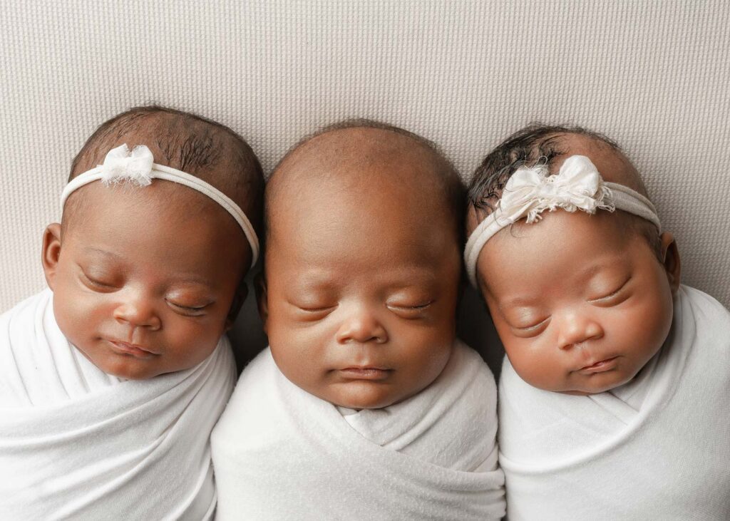 Close-up of three sleeping newborn triplets wrapped in white and resting cheek to cheek.