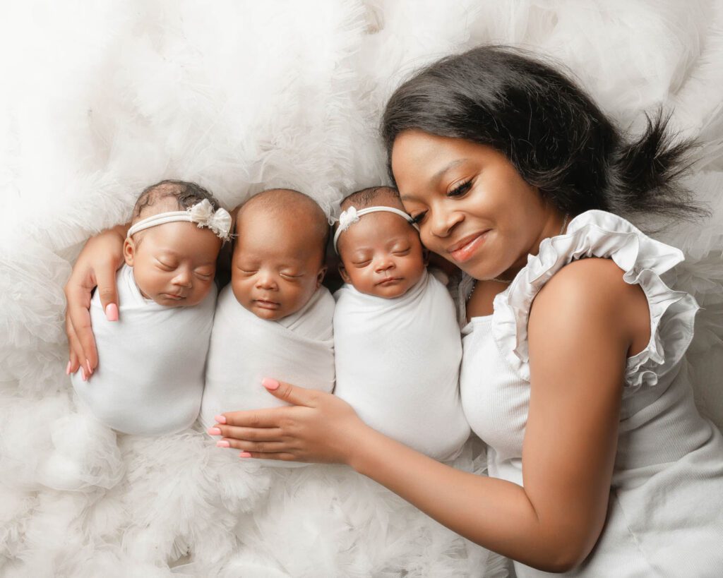 Mom lying beside her three swaddled newborn babies wrapped in white on a soft white fur blanket.