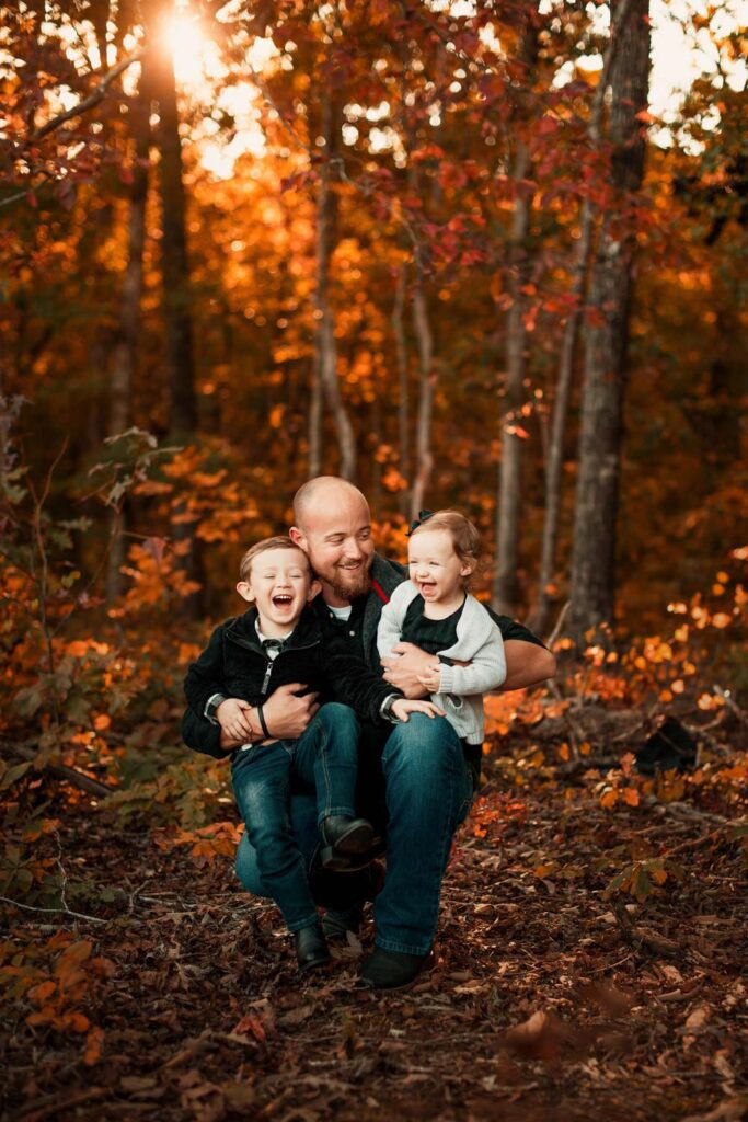 Father kneeling on the ground holding his two kids on his lap as he tickles them while in the forest with red fall leaves. 