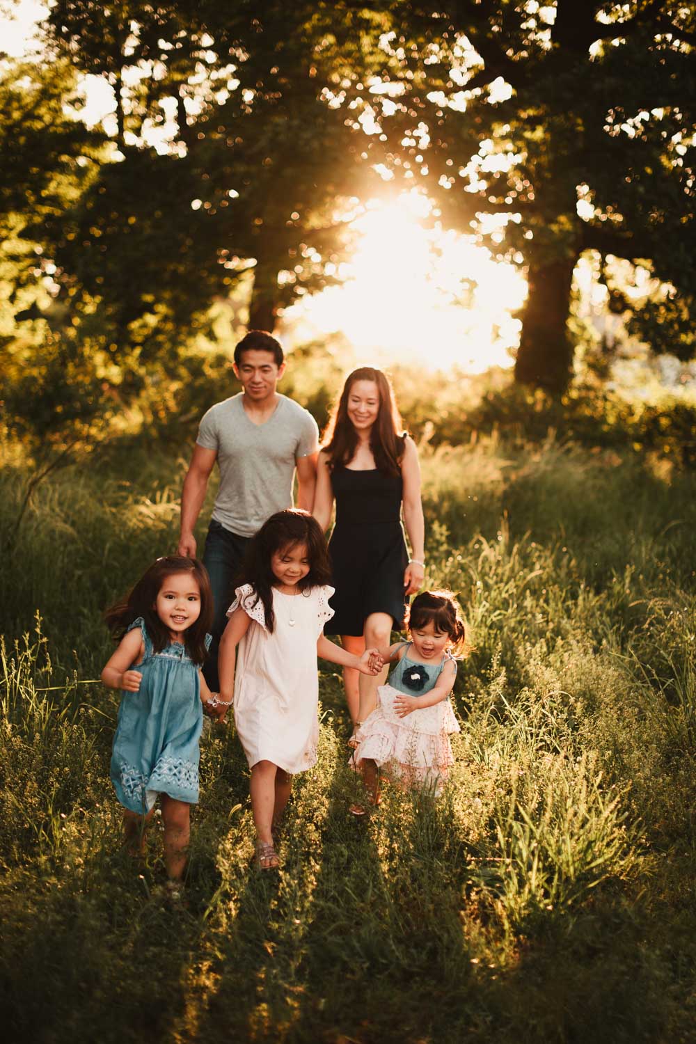Family walking through grass as three daughters hold hands and walk in front of their parents in the golden hour evening.