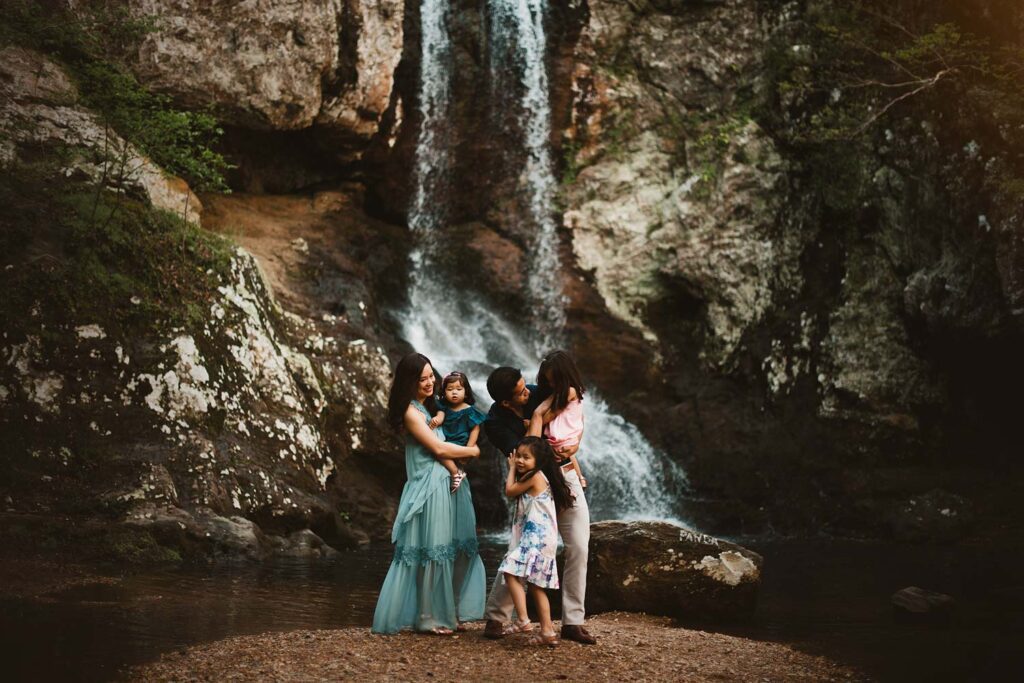 Family of five standing on an island in a pool of water in front of a waterfall basin with soft light surrounding them.