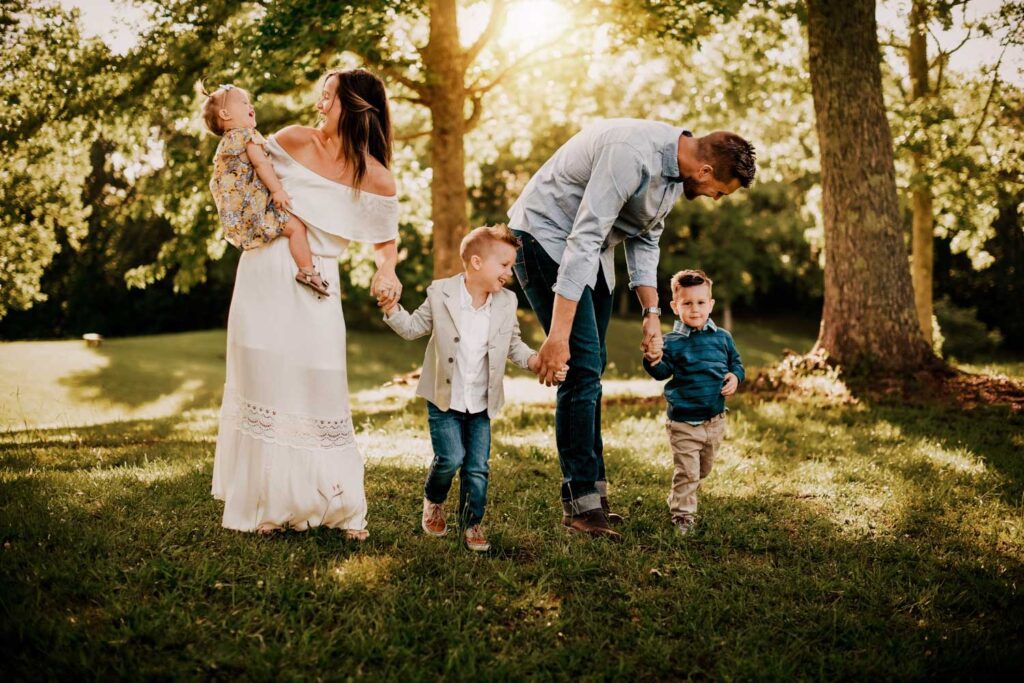Family of five holding hands and walking together in a field of grass with trees behind while wearing blue and white. 