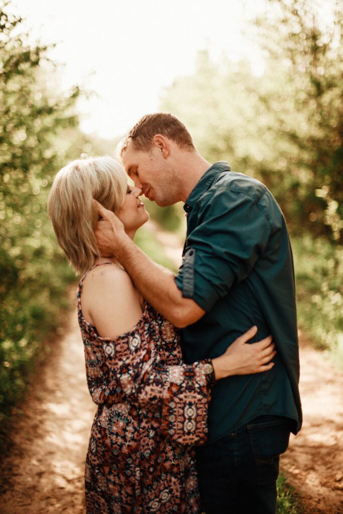 Mom and dad kissing on a wooded path with warm sunlight filtering through the trees.