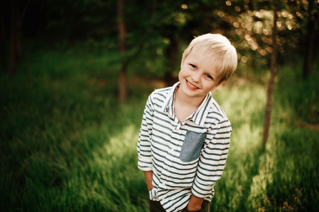 Smiling boy in a striped shirt standing in tall grass with a softly blurred wooded background.