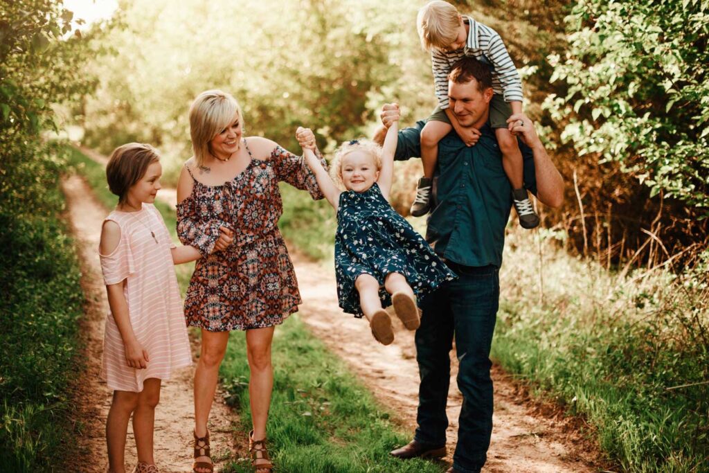 Parents playing with their three children along a grassy trail, with one son sitting on dad’s shoulders.