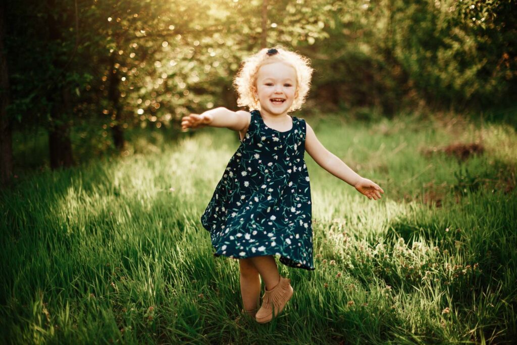 Young girl in a navy floral dress running through tall grass with warm sunlight glowing behind her.
