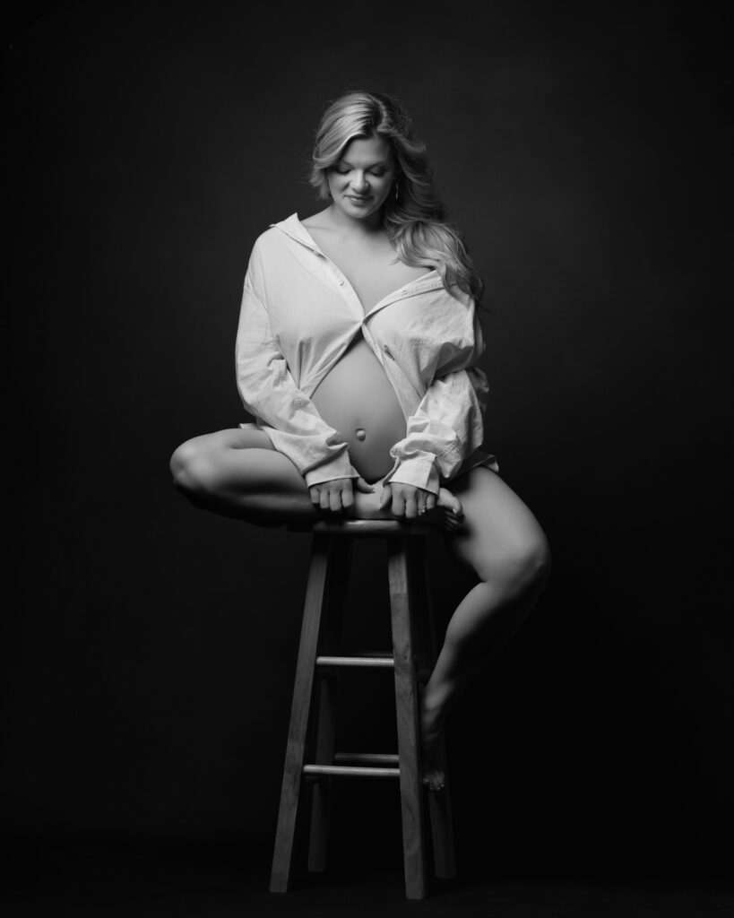 Expecting mother seated on a stool in an open white shirt with her belly exposed.