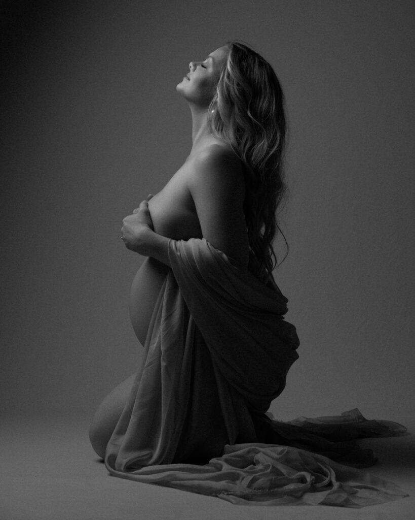 Black and white side-view maternity portrait kneeling with flowing fabric draped around her body.