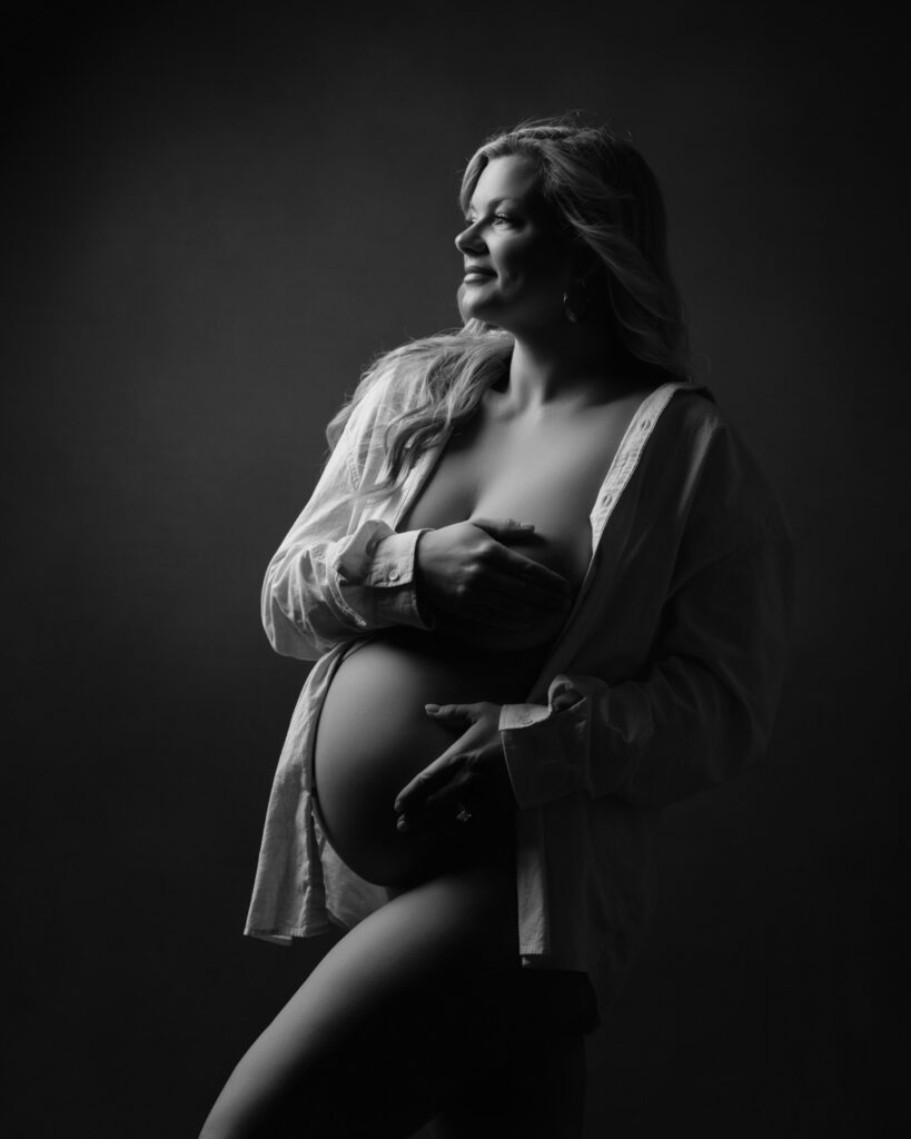 Black and white maternity portrait in an open shirt with both hands resting near her belly.