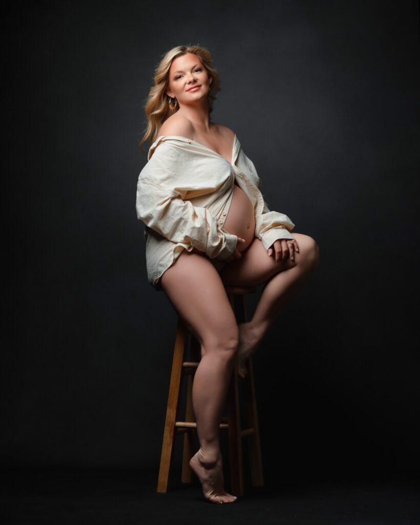 Pregnant mom in open white shirt seated on stool against grey backdrop in studio.