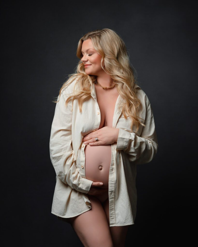 Mom-to-be in an open white shirt cradling her belly against a dark studio backdrop.