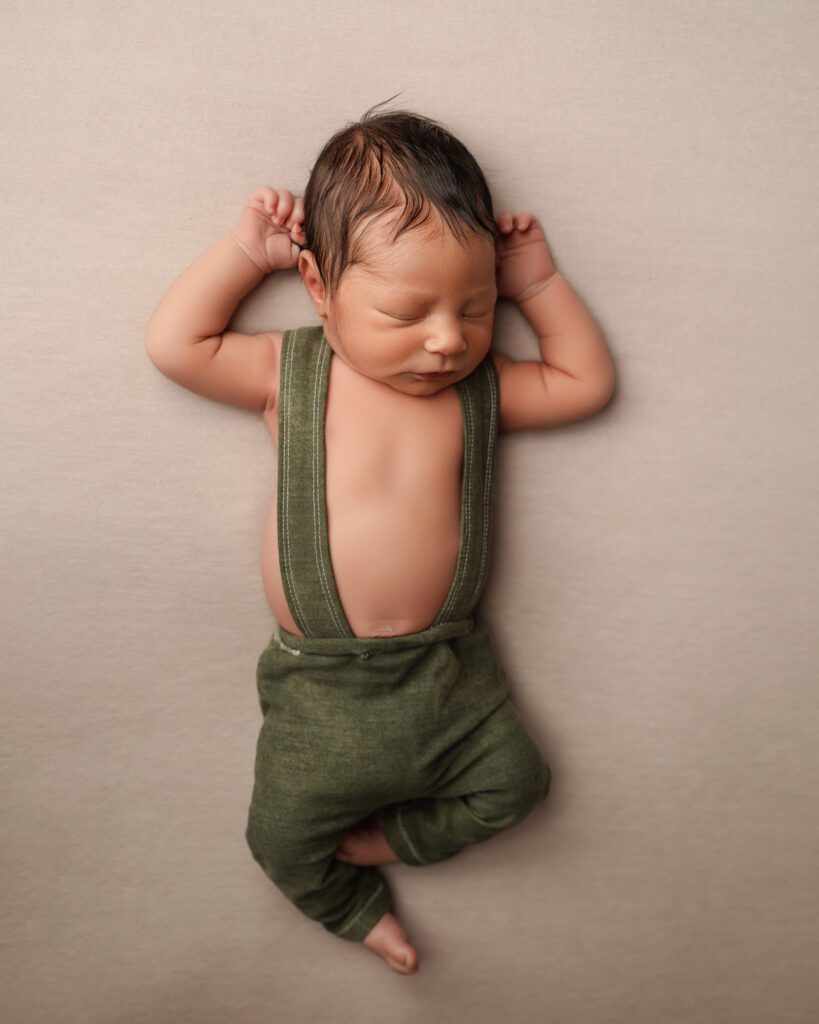 Sleeping newborn in a green suspender outfit with both hands resting near the head on neutral background.