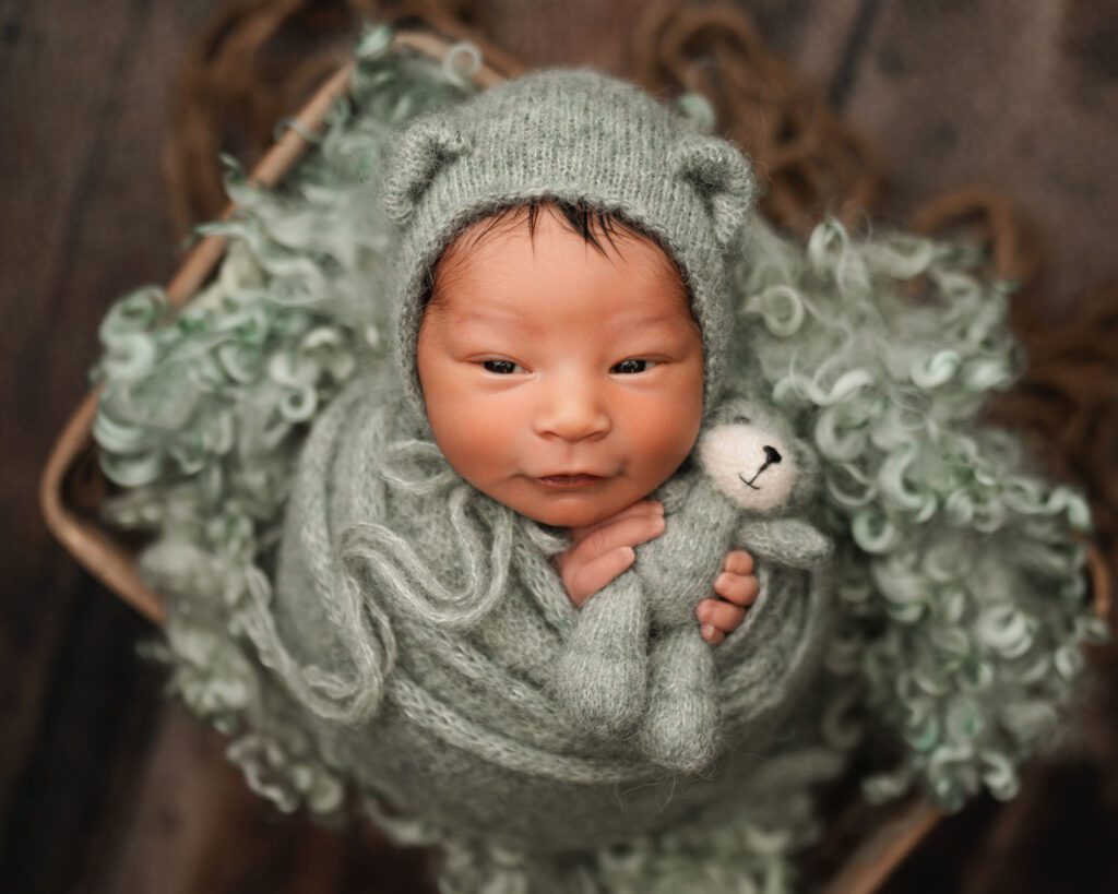 Awake newborn baby wrapped in sage knit and holding a tiny stuffed bear