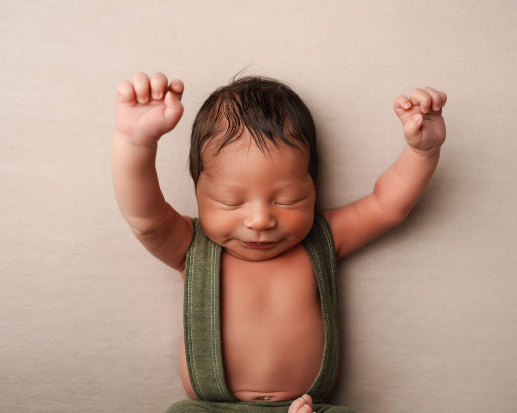 Newborn baby boy wearing green suspender pants stretching his arms up in the air during newborn photoshoot near Buckhead GA