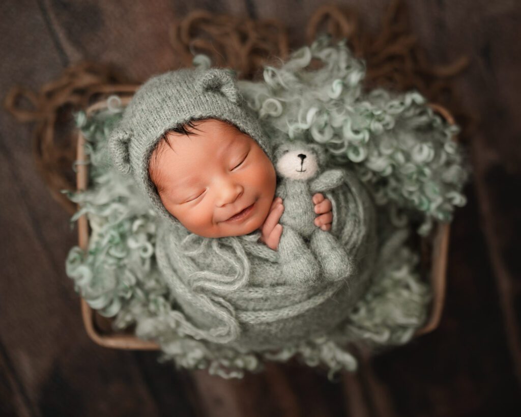 Smiling newborn holding a bear wearing bear bonnet in a wooden basket photographed by Buckhead newborn photographer.
