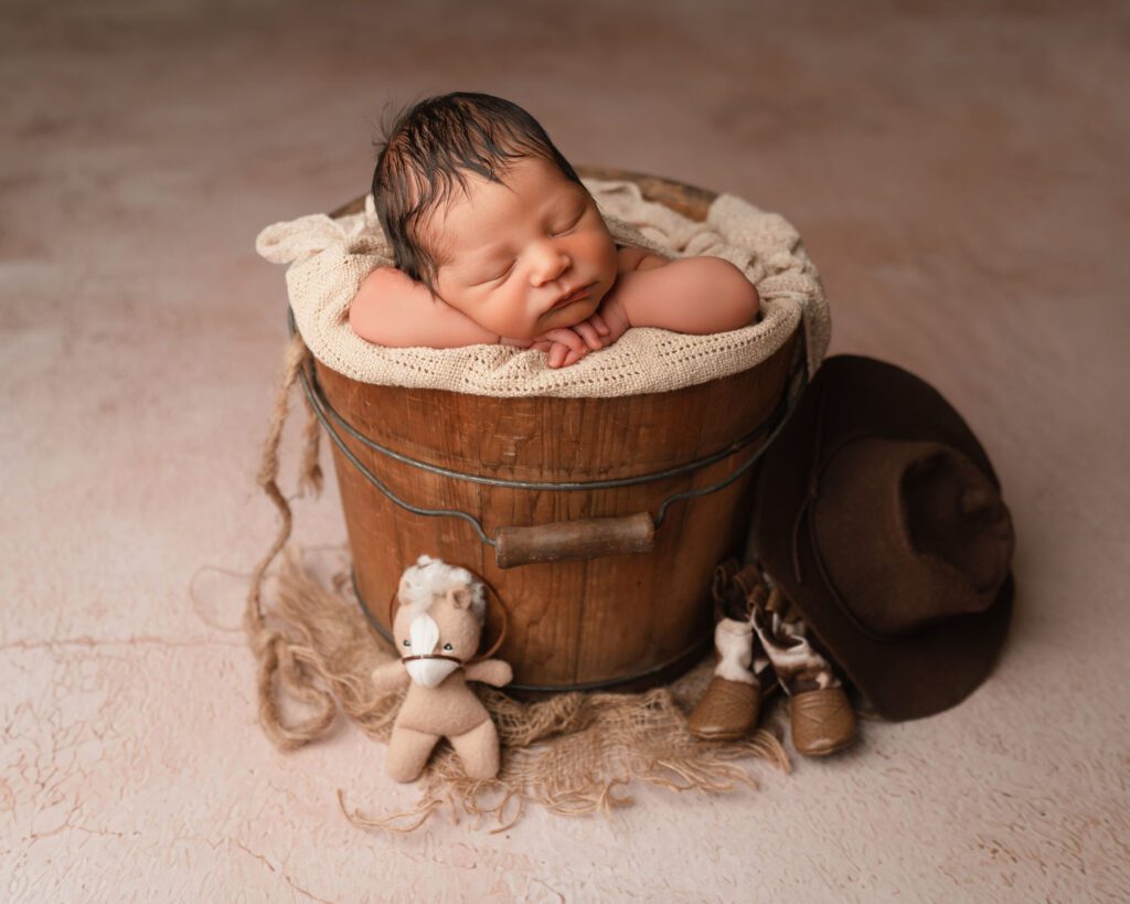 Sleeping newborn posed in a wooden bucket with a cowboy hat and small boots props