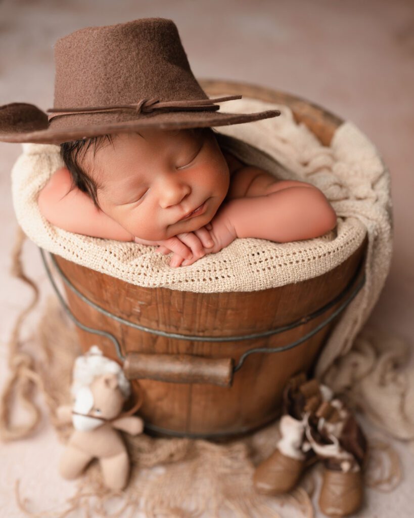 Newborn baby boy sleeping in a wooden bucket wearing cowboy hat with horse and cowboy boot props.