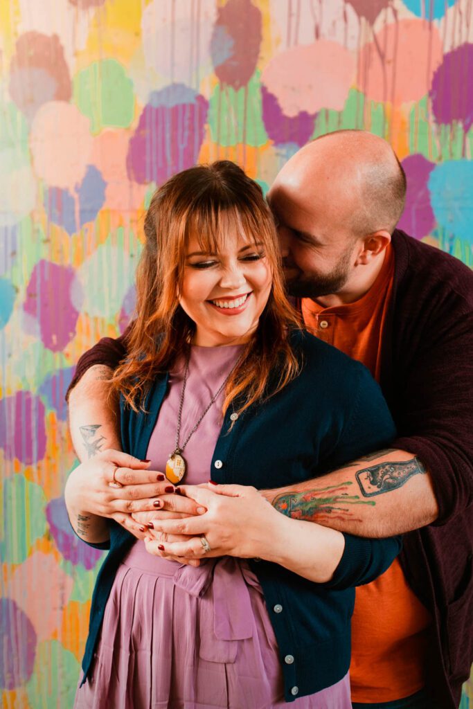 Dad hugging mom from behind as they smile together against Confetti Mural by HENSE.