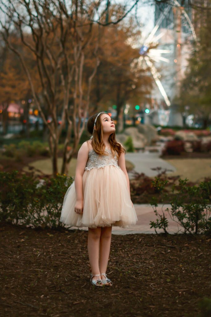 tween girl in cream tulle dress and headband looking up to the sky on front of ferris wheel