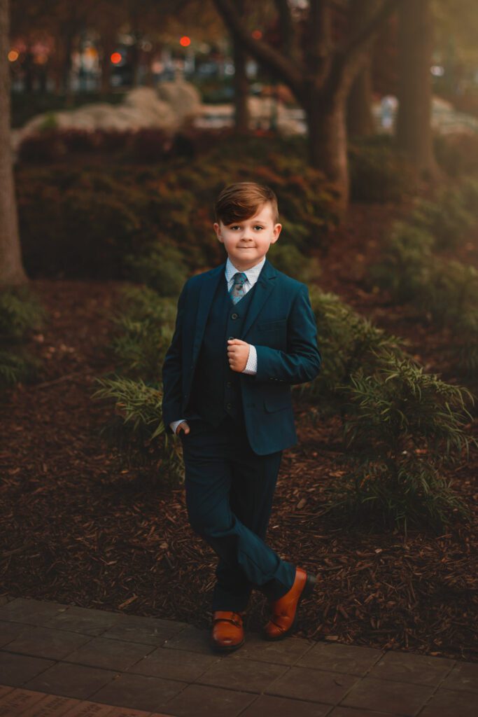 young boy wearing blue suit and tie poses with his hands in his pocket in front of trees at sunset