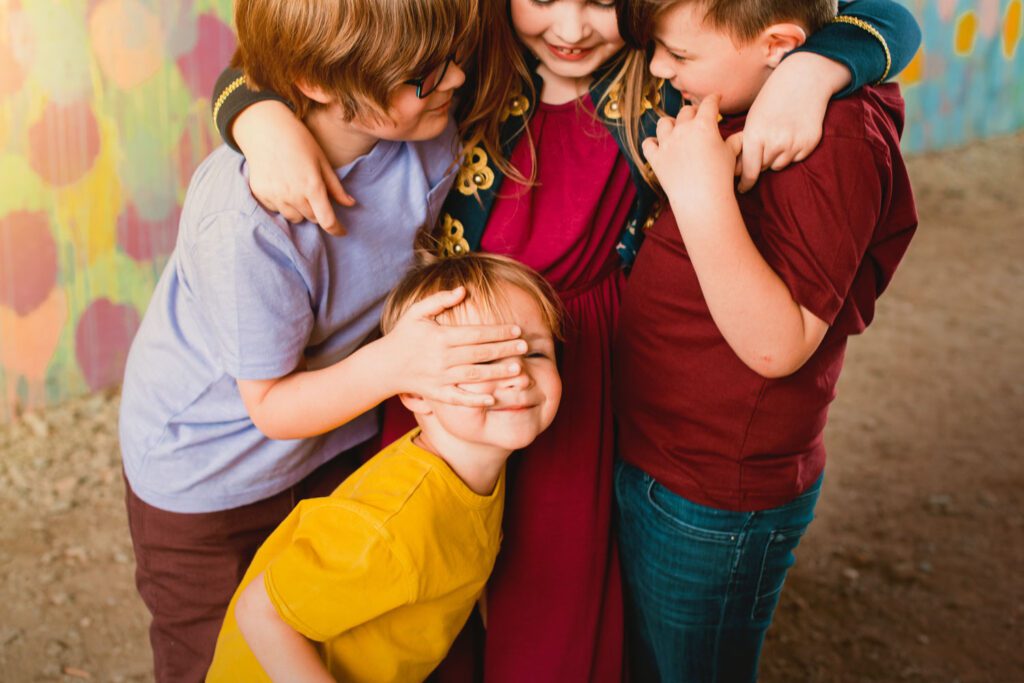 Four siblings huddling together as the youngest covers his eyes in front of a colorful mural.