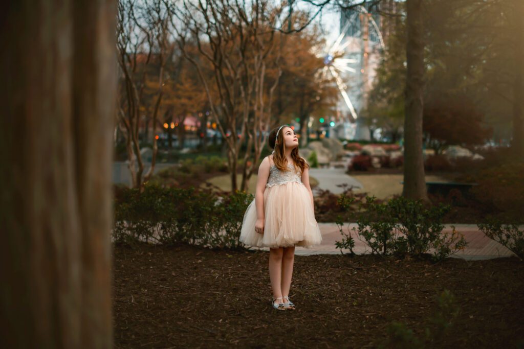 young girl looks at the sky at sunset in sparkly dress outdoors with the Skyview ferris wheel in the background.