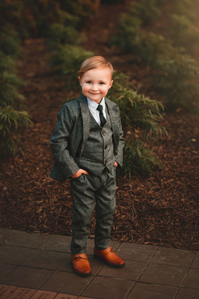 smiling young boy in tailored grey suit, hands in pockets standing on a cement path near greenery.