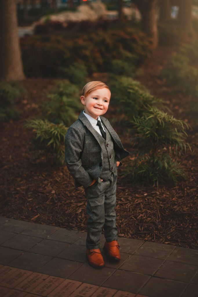 Young boy in a gray suit with hands in his pockets standing on path among landscaped greenery.